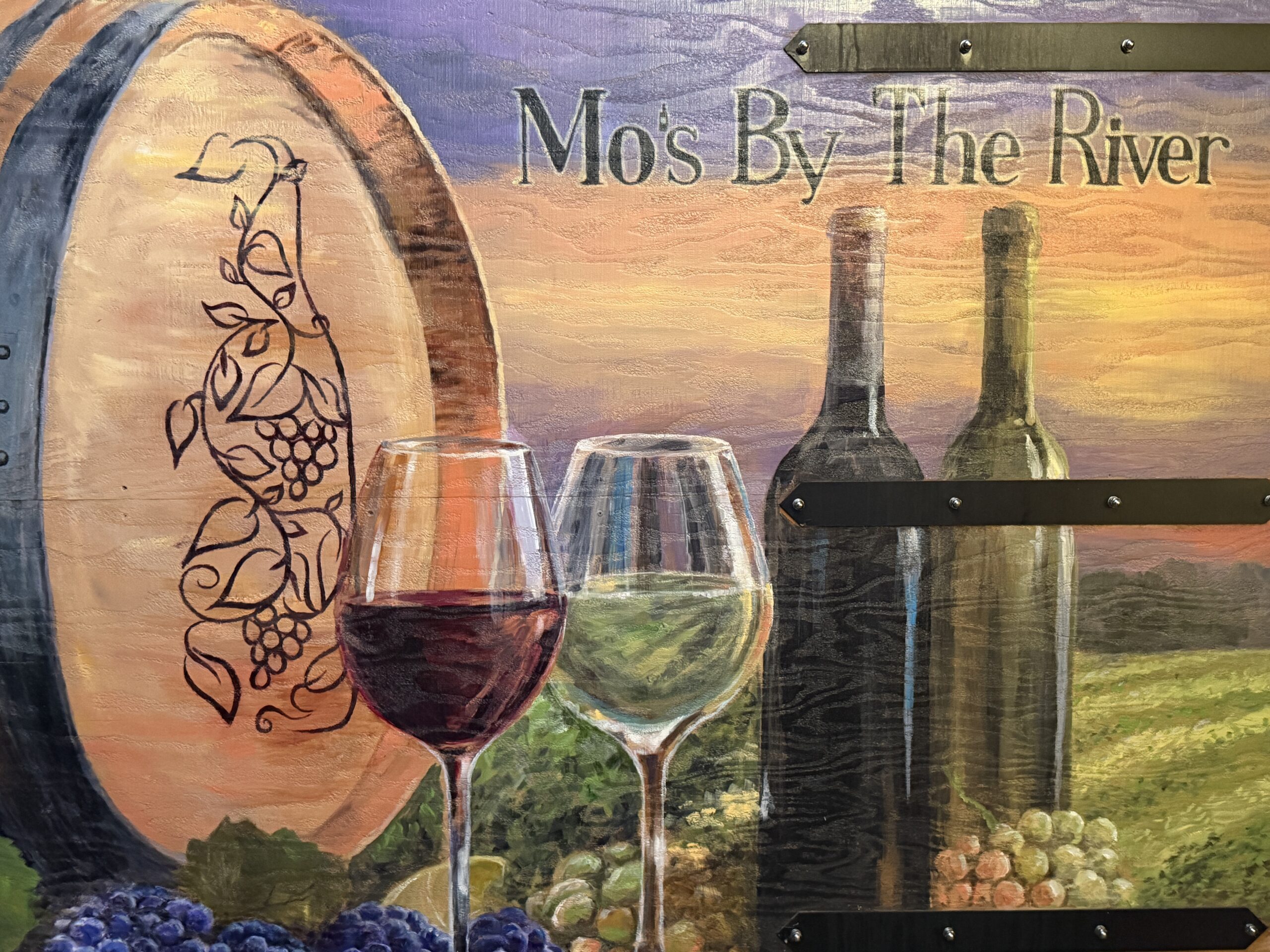 A hand-painted sign reading Mo's By The River, with a wine barrel, a glass of red and a glass of white, and two bottles set against a sunset over vineyards, painted on wood planks held by black iron straps.
