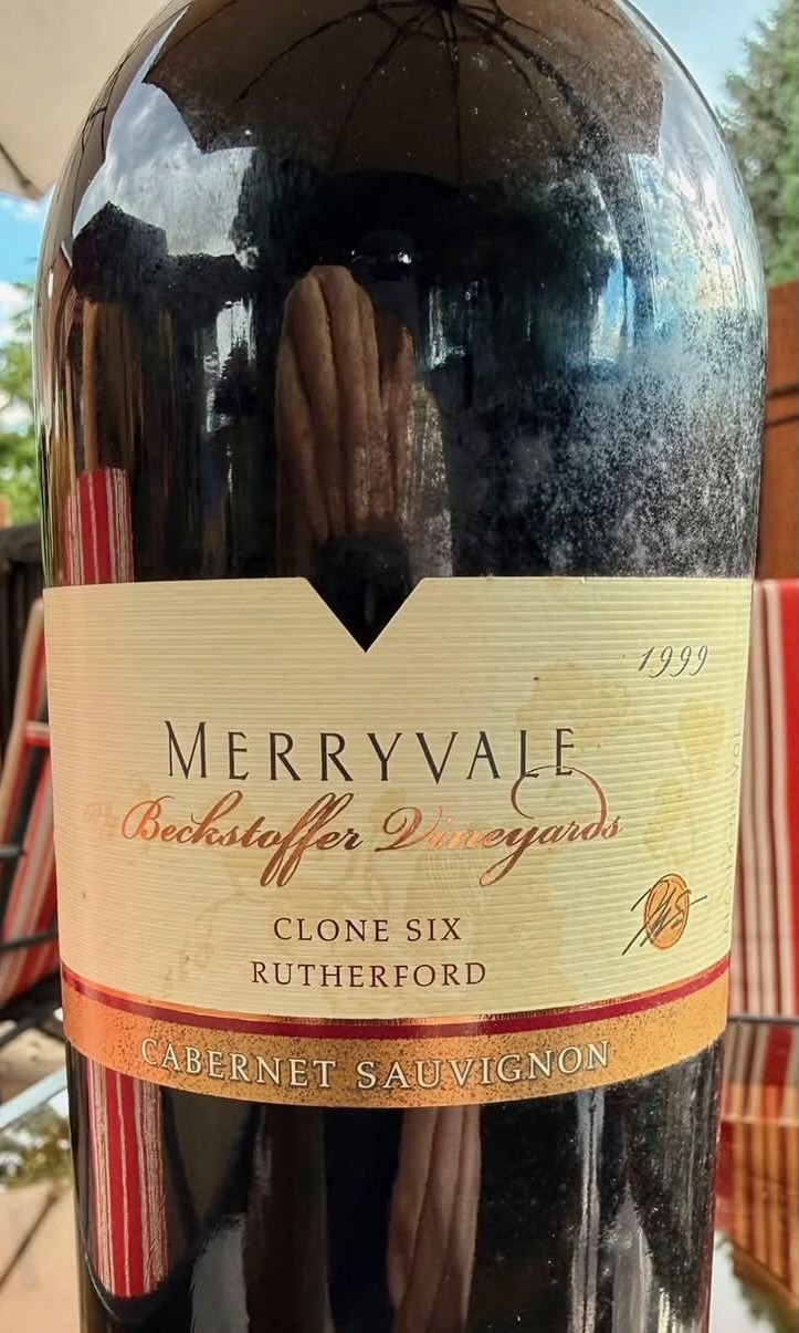 A dusty bottle of 1999 Merryvale Beckstoffer Vineyards Clone Six Rutherford Cabernet Sauvignon on the patio at Mo's by the River, with a patio umbrella and striped chairs reflected in the dark glass.