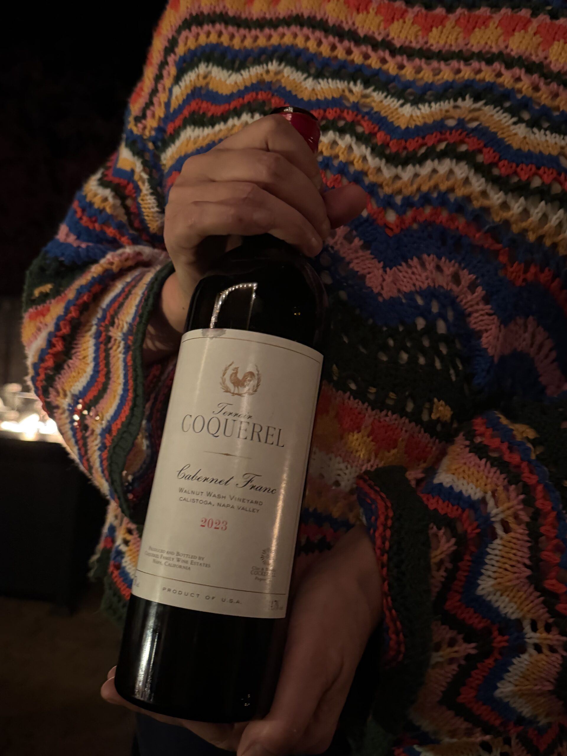 Mo, in a brightly striped knit sweater, holds a bottle of 2023 Terroir Coquerel Cabernet Franc from Walnut Wash Vineyard, Calistoga, Napa Valley. The white label has a gold rooster crest, and candlelight glows in the dark background.