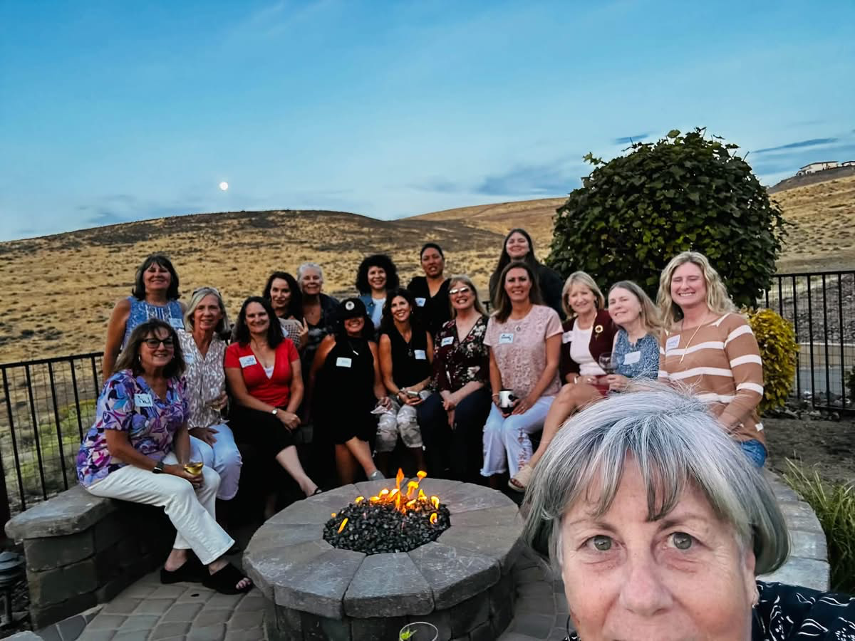 A group of about seventeen women wearing name tags gathers around a stone fire pit on a backyard patio at dusk, many holding wine glasses. Dry sagebrush hills and a rising moon are behind them, and a gray-haired woman taking the selfie fills the lower right corner.