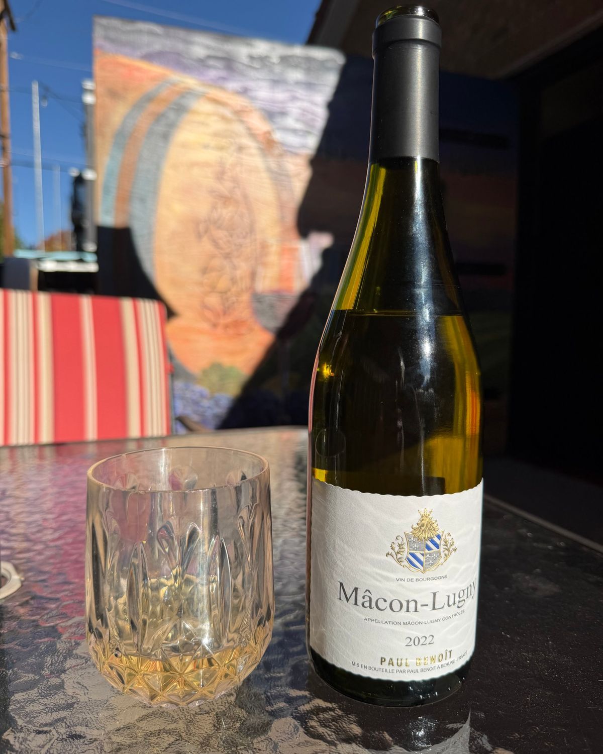 A bottle of Paul Benoît Mâcon-Lugny 2022 beside a cut-glass tumbler of white wine on a sunny patio table, with a painted wine barrel mural behind it.