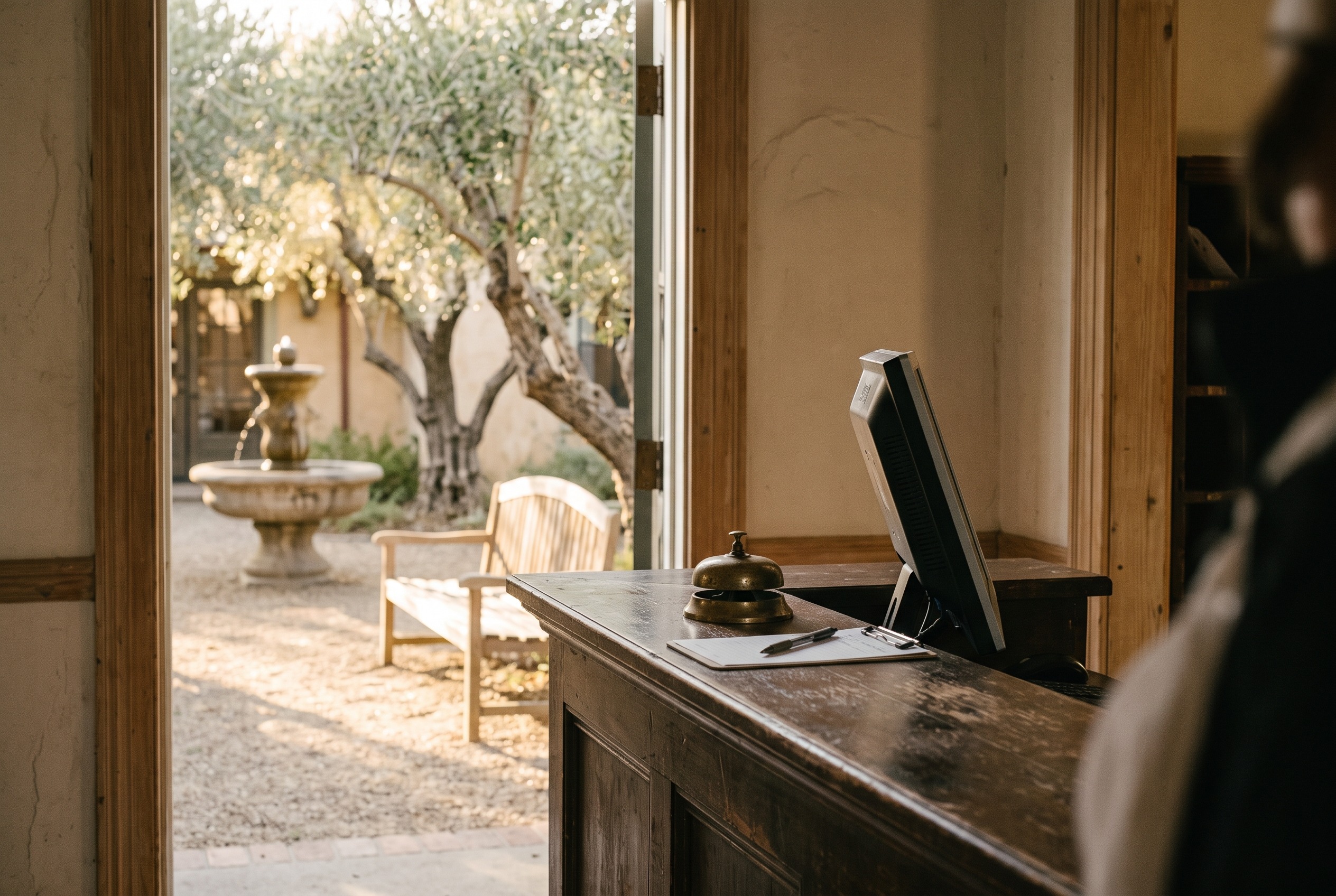 Late afternoon light at the front desk of a small Calistoga inn, with old olive trees and a stone fountain visible through the open door to the courtyard.